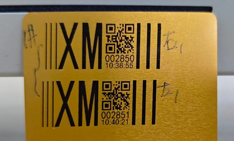 A test print on a brushed aluminium sheet: the XinmoJet wordmark, a 2D code and a six-digit serial number in black solvent ink