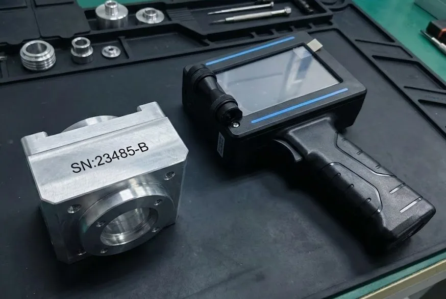 A handheld TIJ printer standing beside a machined aluminium part carrying a black solvent-ink serial number